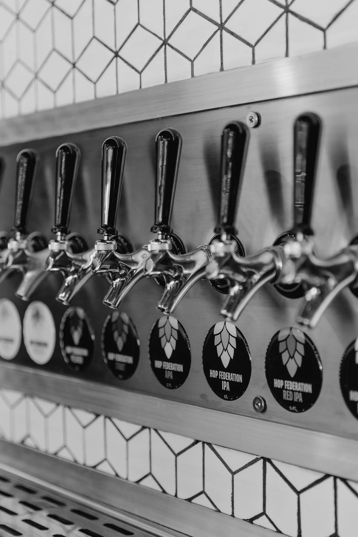A black and white photo of a wall with a metal rack holding several tap handles for beer pouring, with labels for different beer types including 'Hop Federation IPA' and 'Hop Federation Red IPA'.