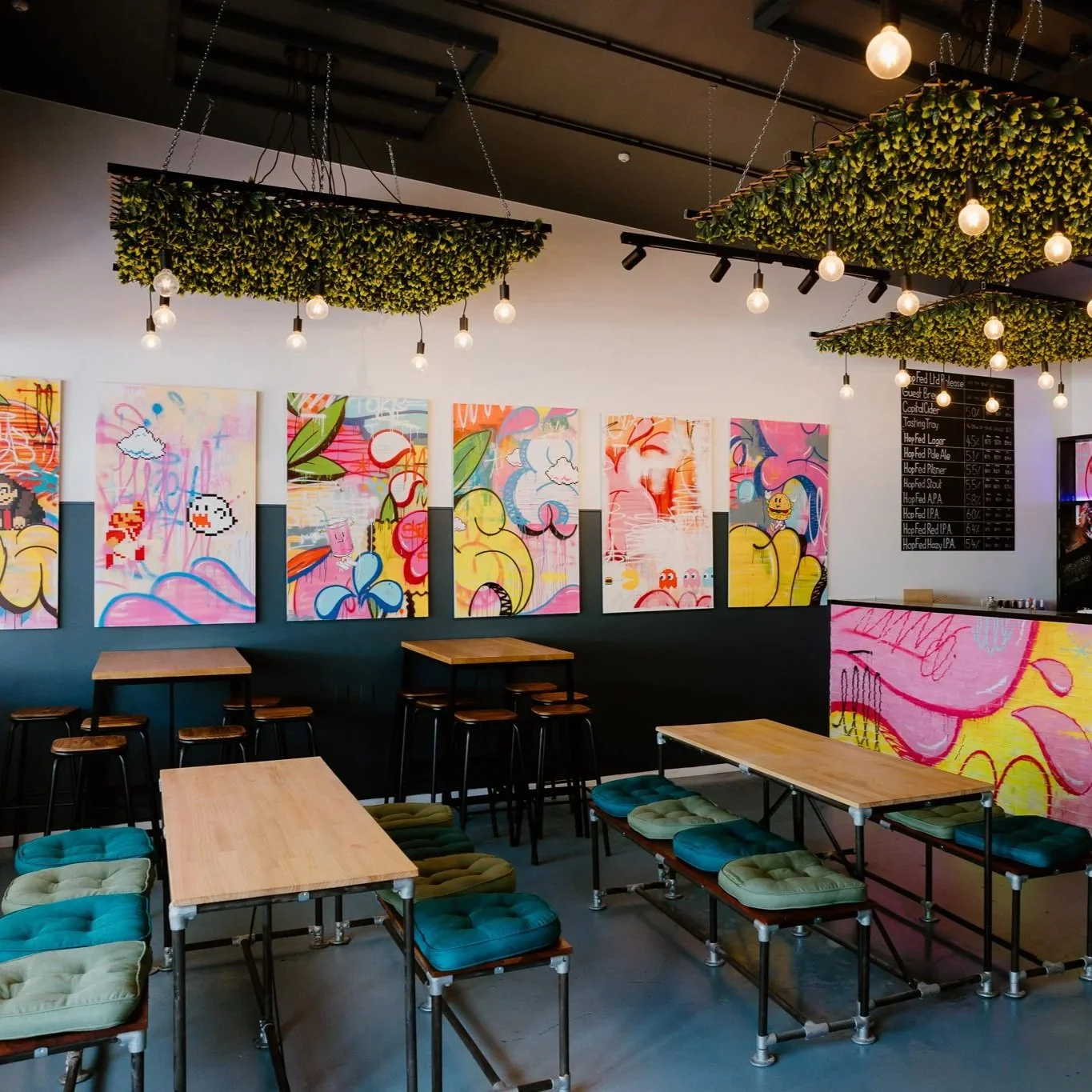 A modern cafe with colorful abstract and pixel art-style paintings on the wall, wooden tables with cushioned stools, and decorative ceiling installations of greenery with hanging light bulbs.