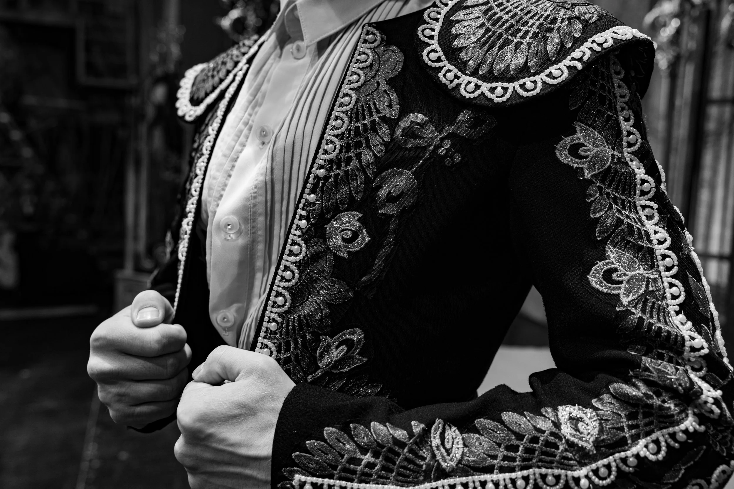 Vue en gros plan d'une personne portant une veste noire avec des broderies et des perles, et une chemise à rayures blanches et noires. La personne a ses mains en poing, leur position suggère une posture de confiance ou de défi.