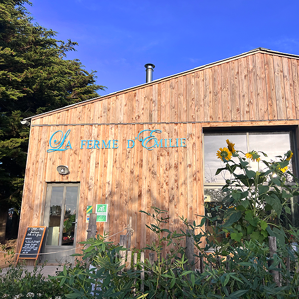 Façade en bois d'une ferme avec un panneau indiquant 'La Ferme d'Emile', des fleurs de tournesol et des affiches.