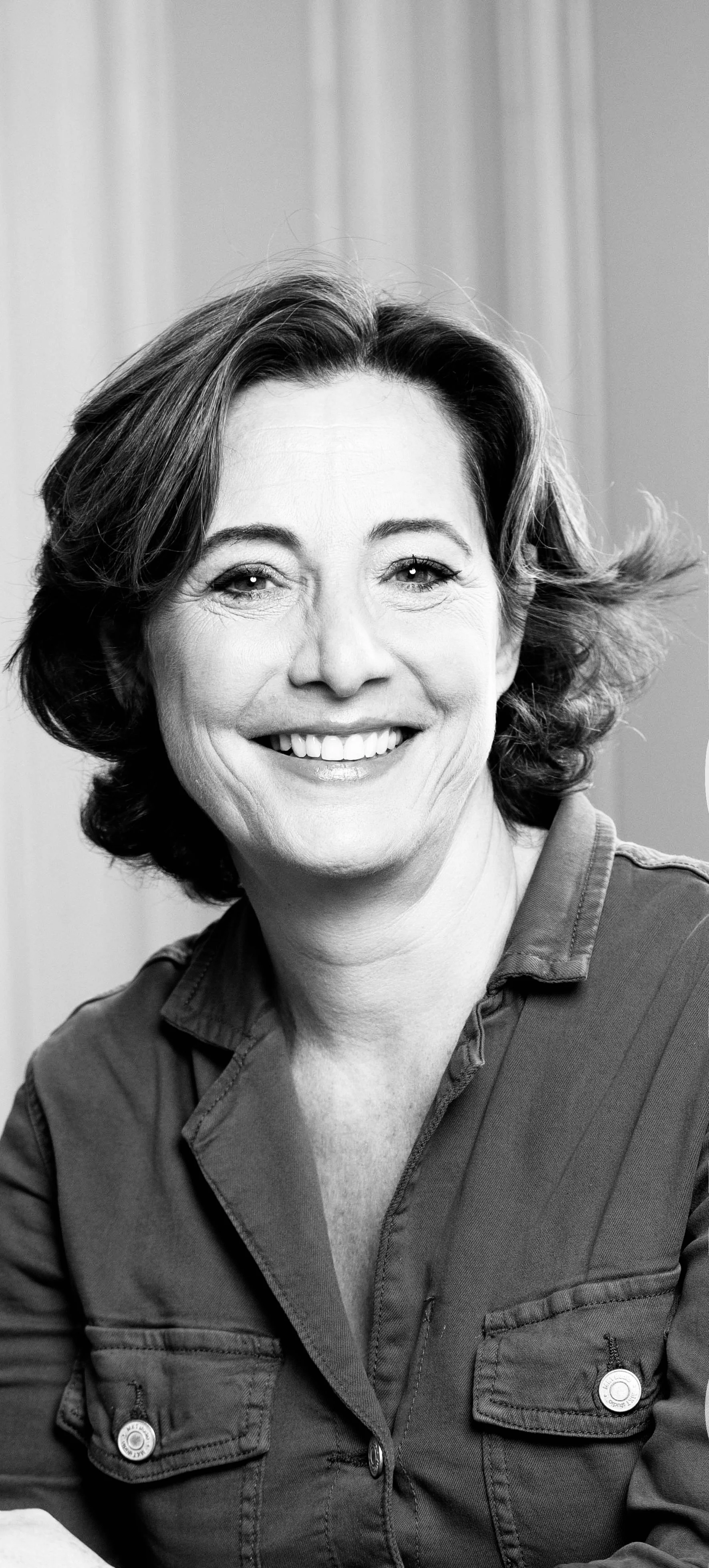 Portrait en noir et blanc d'une femme souriante, yeux brillants, cheveux courts et bouclés, vêtue d'une veste en coton.