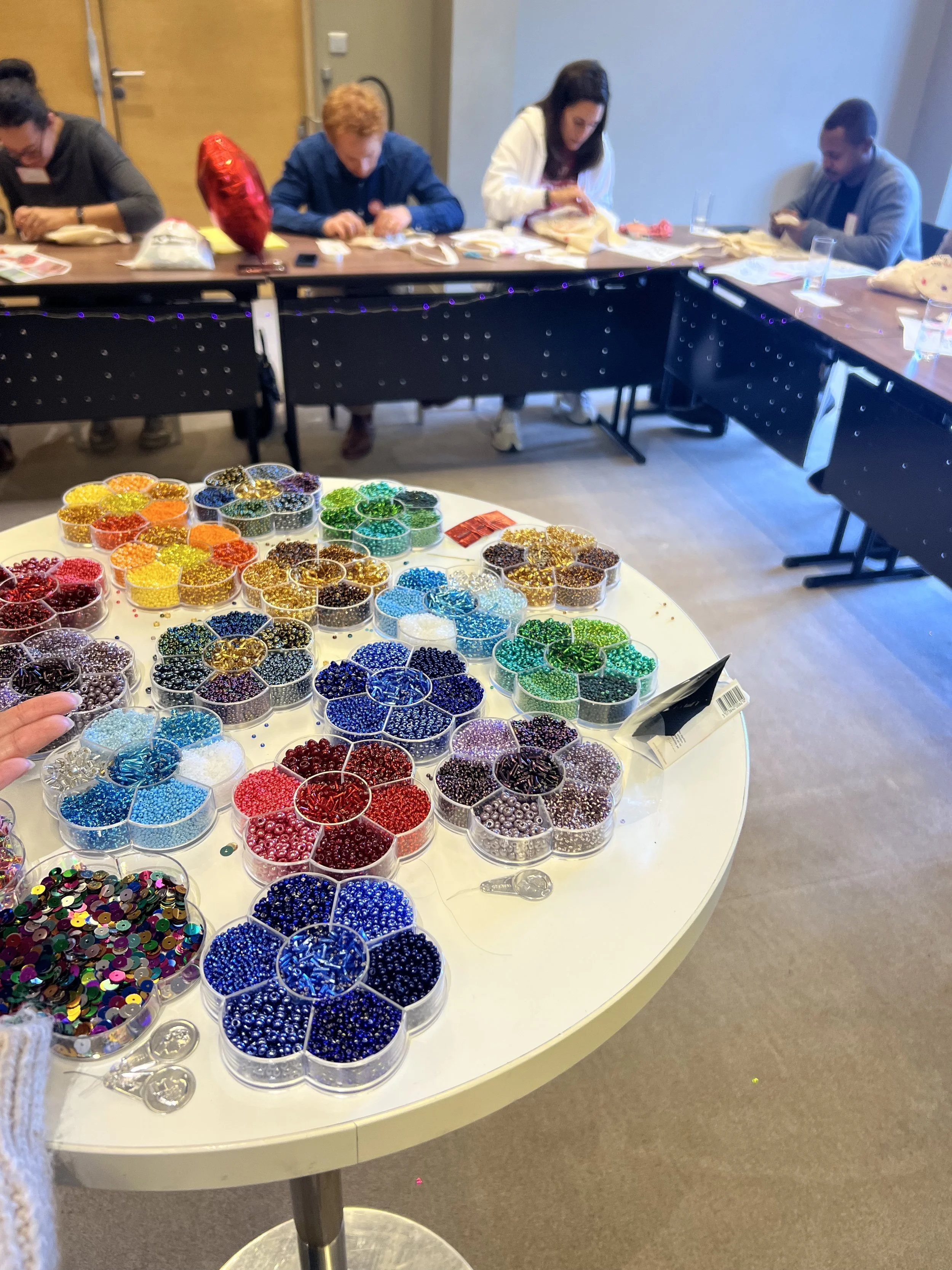 Une table ronde avec de nombreuses boîtes transparentes remplies de perles colorées pour être utilisées dans la création de bijoux, situées dans une pièce avec plusieurs personnes au fond qui travaillent ou trient également des perles.