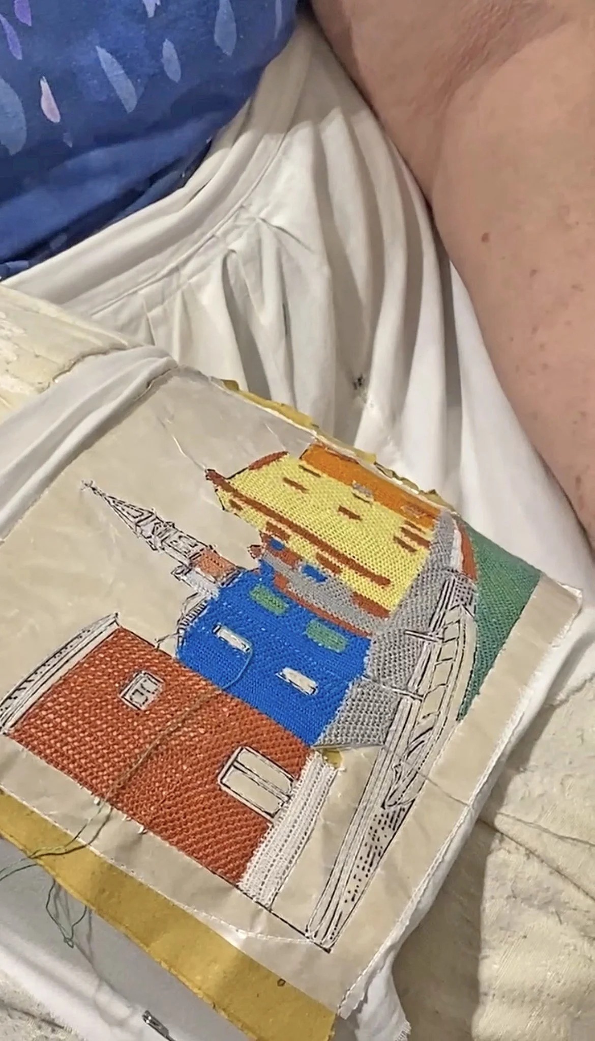 Une broderie en cours représentant une ville avec des bâtiments colorés, sur une toile de coton.