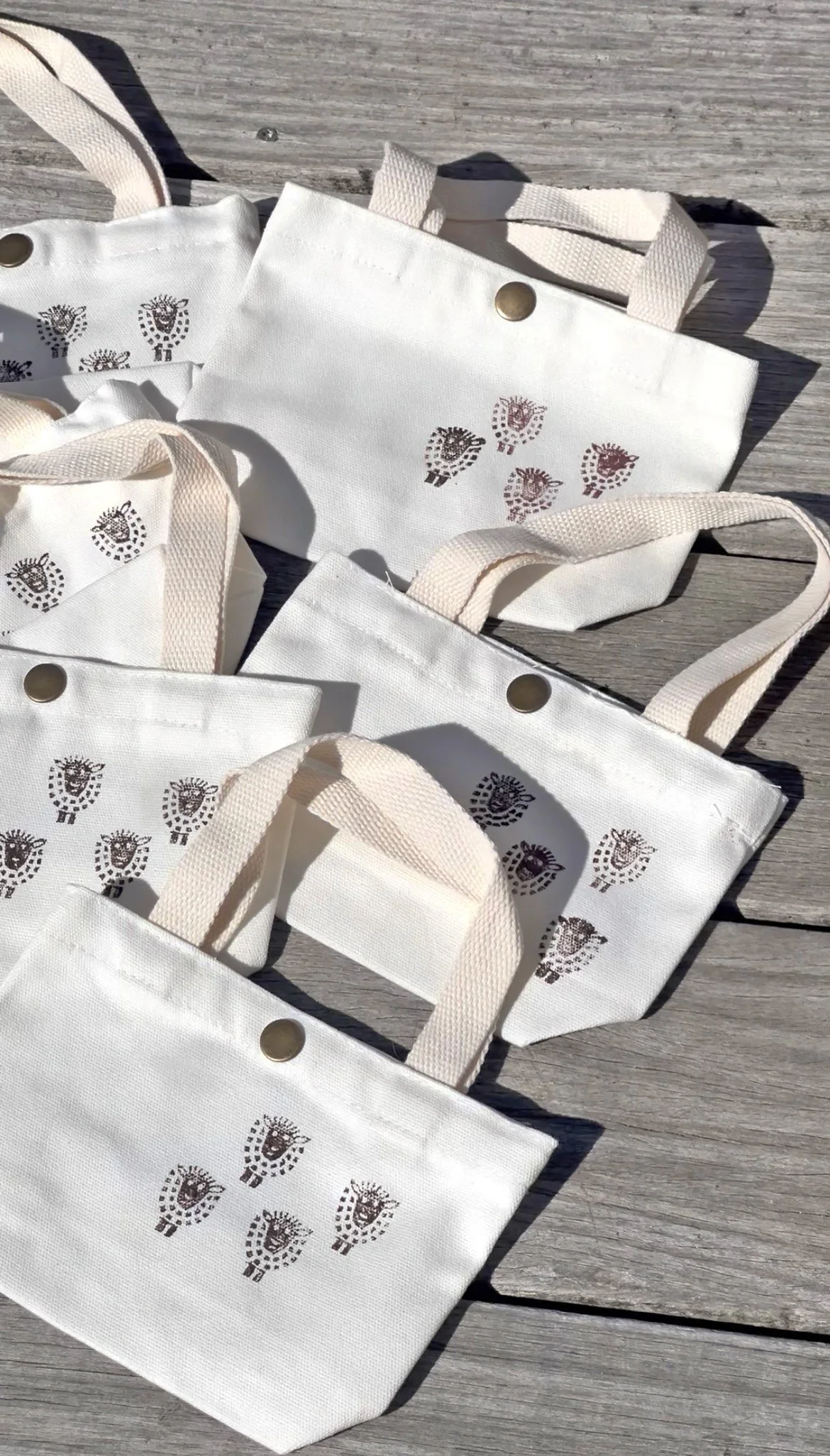 Multiple white tote bags with beige handles and a printed leaf design lie on a wooden surface.