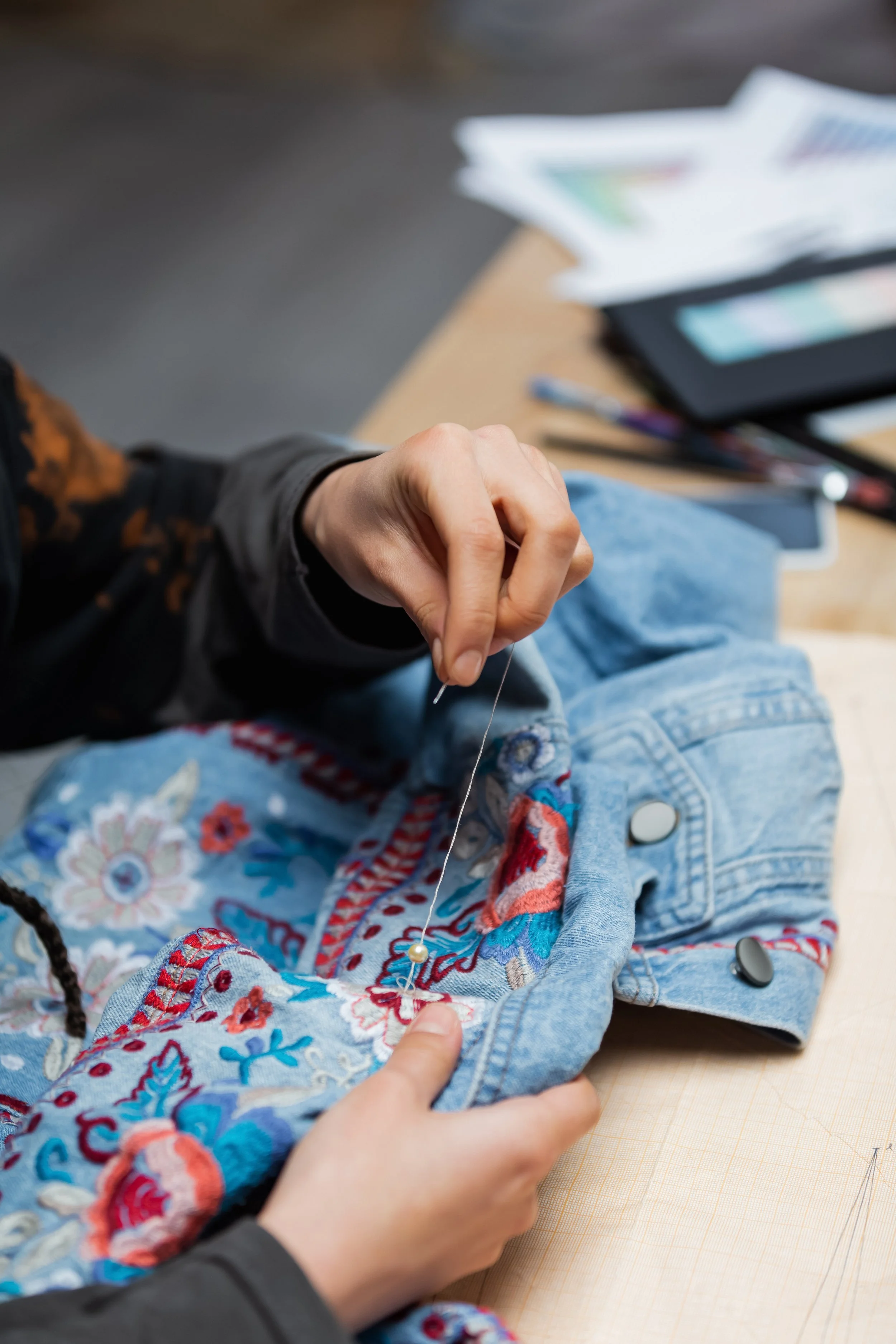 Gros plan d'une main en train de coudre avec une aiguille dans un vêtement en jean à broderie colorée posé sur une table en bois, avec une tablette, un stylo, et des papiers en arrière-plan.