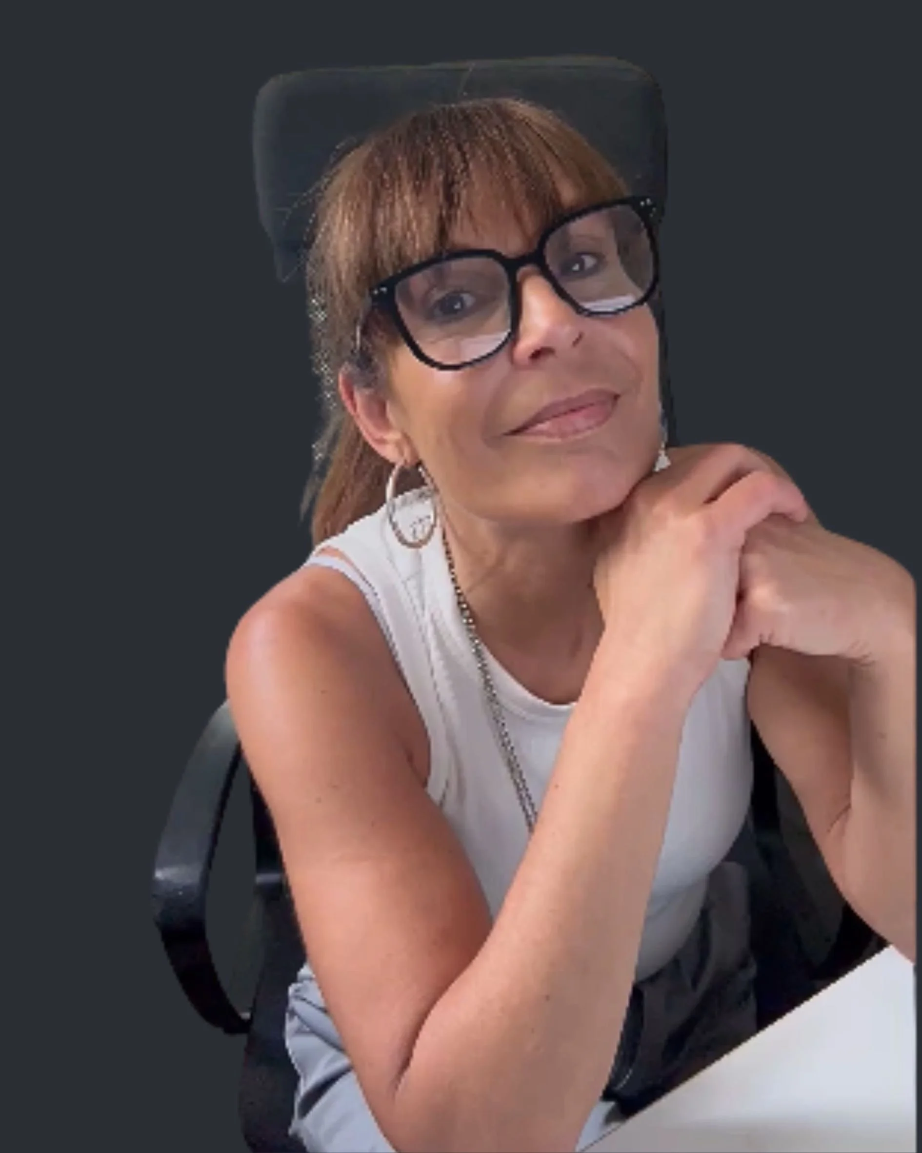 Eine Frau mit braunen Haaren, Brille und Ohrringen lächelt, während sie an einem Tisch sitzt und ihre Hände unterhalb ihres Gesichts hält.