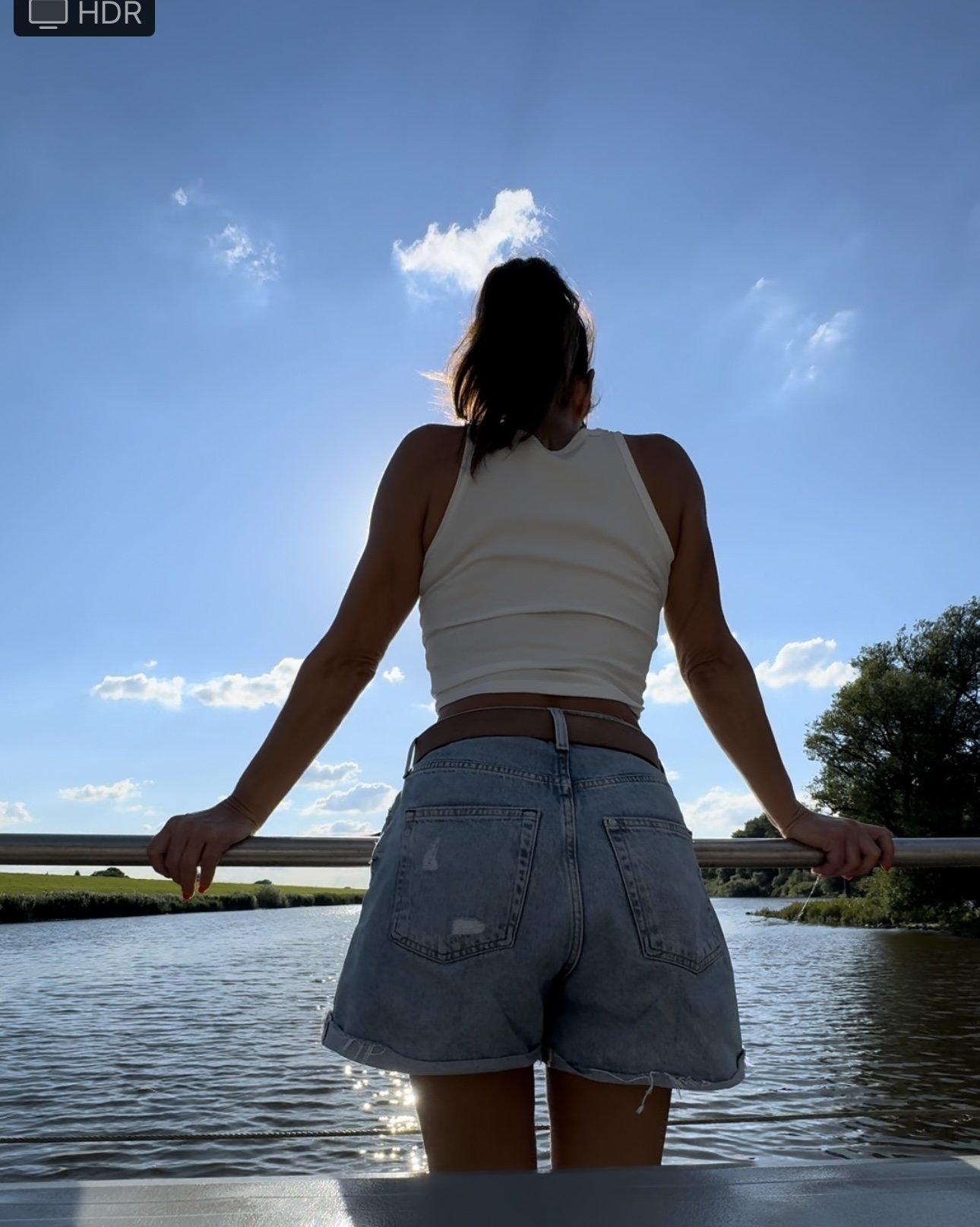 Junge Frau steht am Flussufer, betrachtet die Wasserlinie bei Sonnenschein, trägt weiße Top und Jeansshorts, umgeben von Natur, mit klarer blauer Himmel und wenigen Wolken.