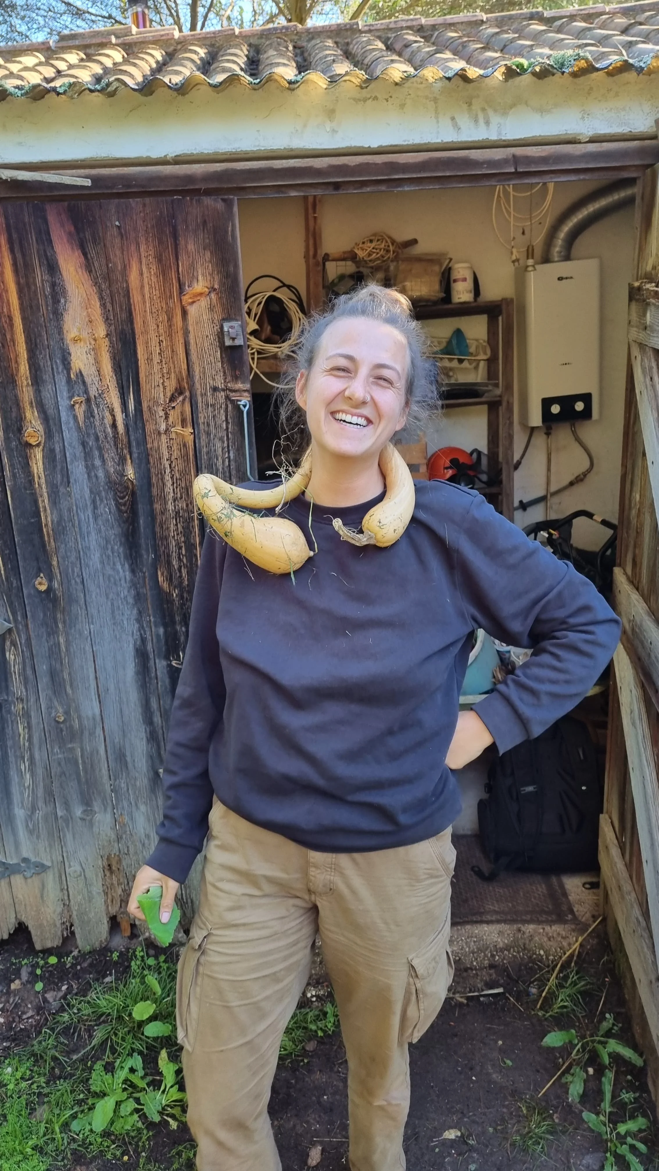 Vrouw met een grote gele pompoen rondom haar hals, lachend en poseert in een tuin of werkplaats.