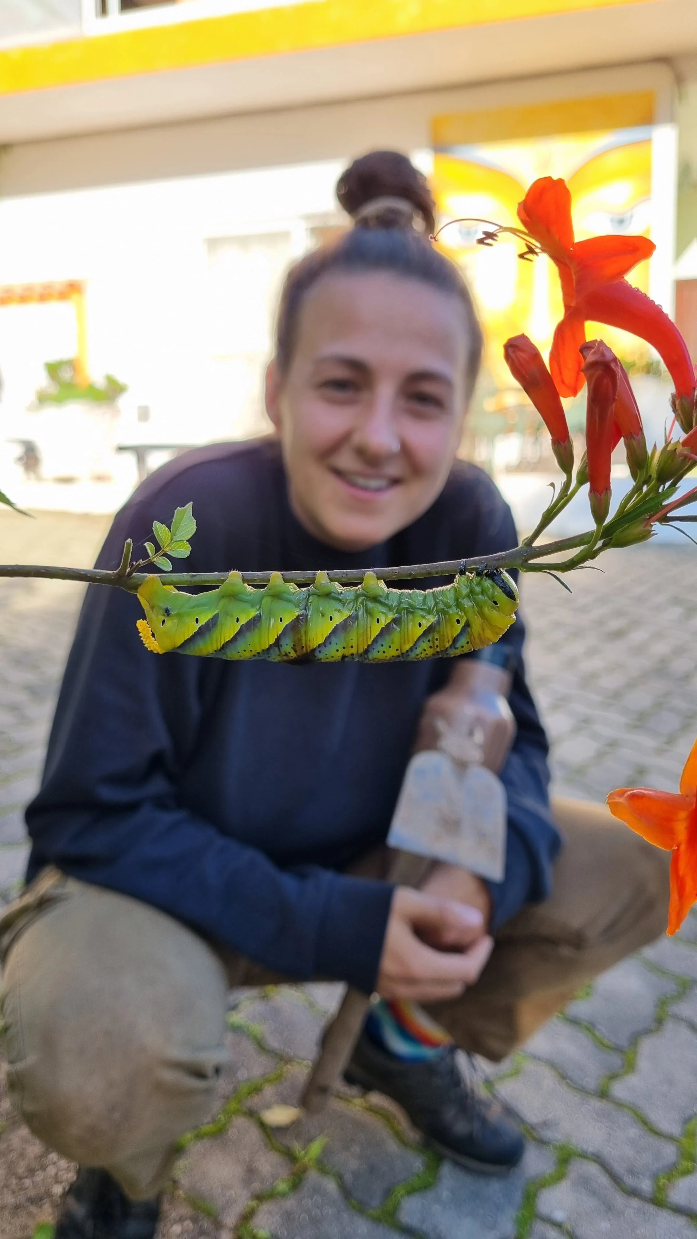 Een grote groene rups met zwarte stippen op een tak, de achtergrond bevat een lachende vrouw met donker haar in een donkere jas, al zoomend wordt de rupseen nietig door de bloem. Ze zitten buiten onder een overkapping met gele en witte details.