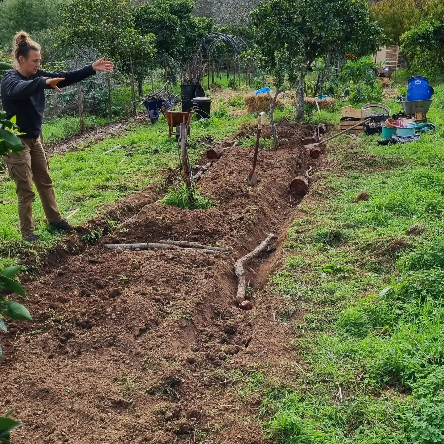 Een persoon graaft een lange sleuf in de tuin met verschillende gereedschappen en tuinspullen op de achtergrond.