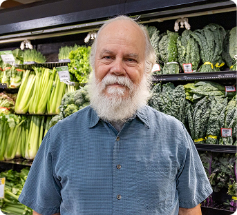 An elderly man with a beard and a blue shirt standing in a grocery store's produce section with various leafy greens and vegetables behind him.