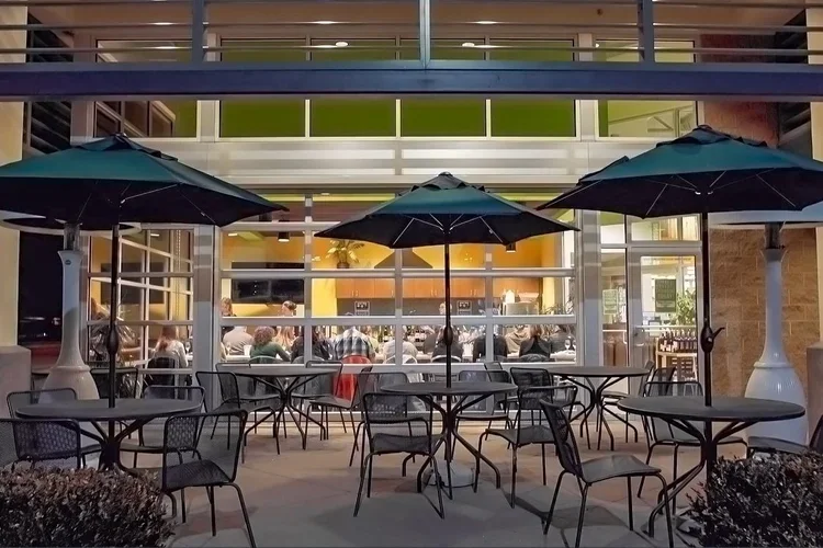 Outdoor patio seating area at a restaurant with tables, chairs, and umbrellas, viewed through large glass windows showing indoor dining.