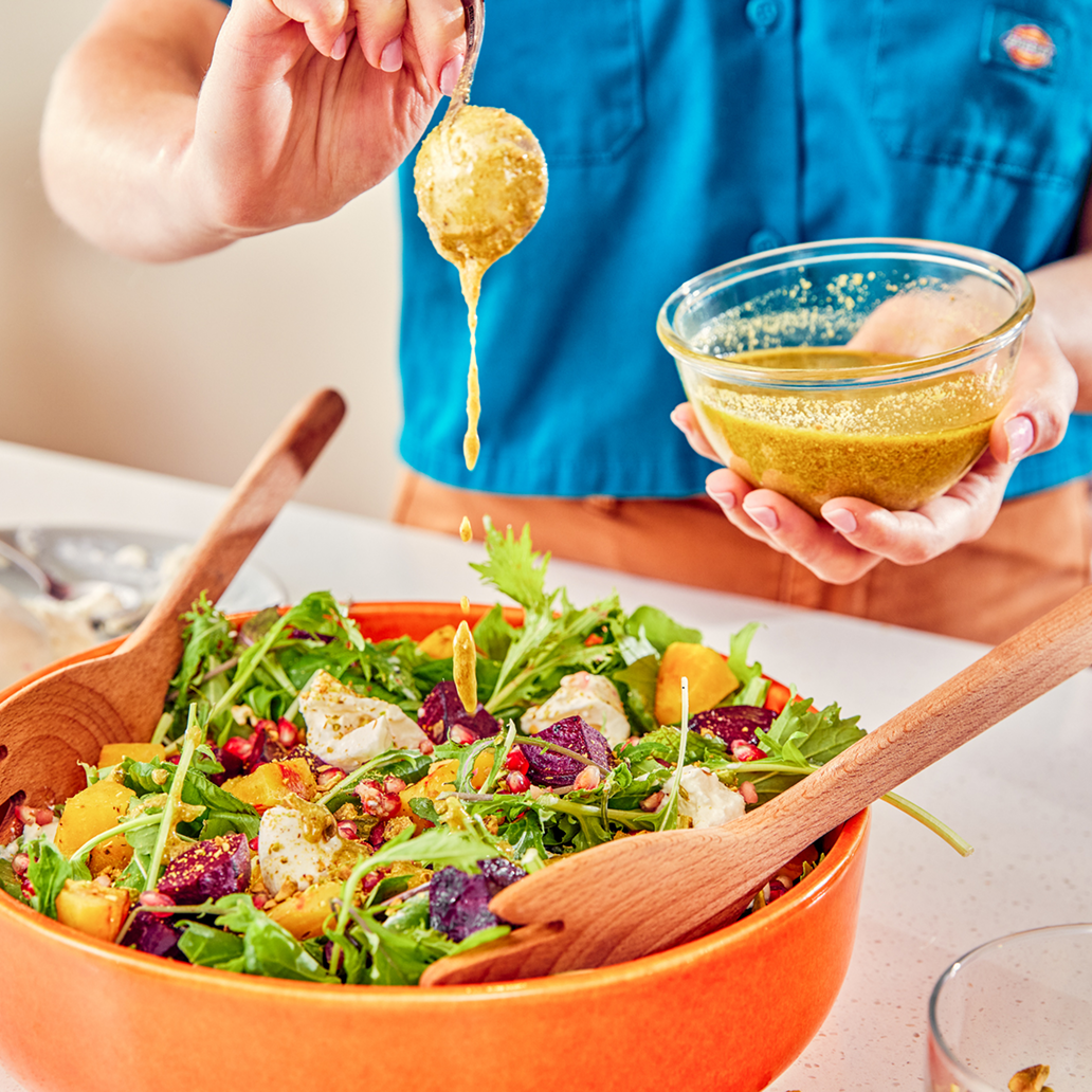 Person in a blue shirt pouring salad dressing from a glass bowl onto a colorful mixed salad in an orange bowl, with salad utensils inside.