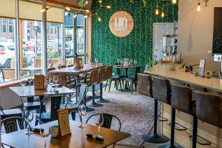 Interior of the Boise Co-op Market cafeteria area with tables set with glasses and menus, bar seating with leather chairs, a green decorative wall with a round sign, and large windows allowing natural light.