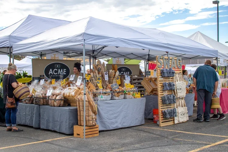 Farmer's market stall selling baked goods and cloth bags, with people shopping and a tent overhead.