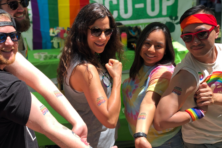 A group of five diverse people at a pride event, showing their arm tattoos with rainbow colors, smiling and flexing their muscles, with a rainbow pride flag and a sign in the background.