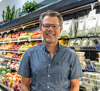 A man with glasses and gray hair smiling in a grocery store aisle filled with fresh fruits and vegetables.
