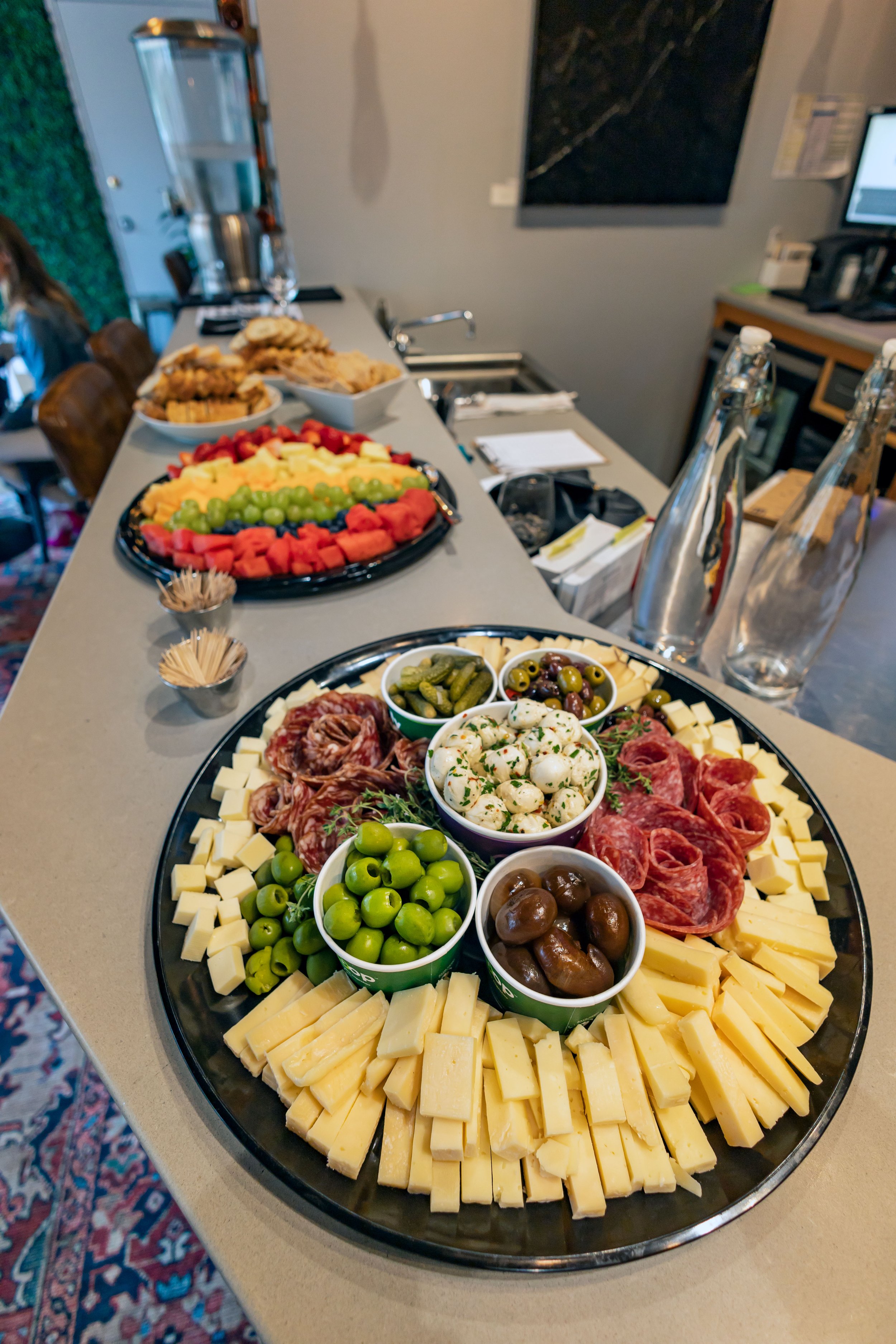 A cheese and charcuterie platter with olives, grapes, and other snacks, along with several bowls of sliced fruits and crackers, is laid out on a long table.