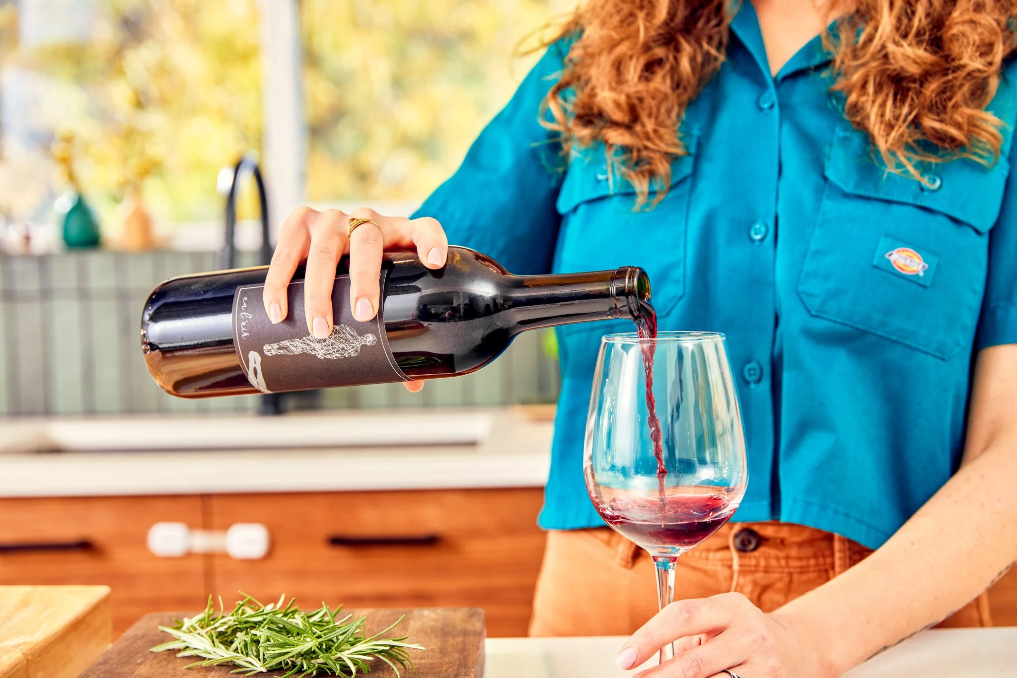 A person with curly red hair wearing a blue shirt is pouring red wine into a glass.