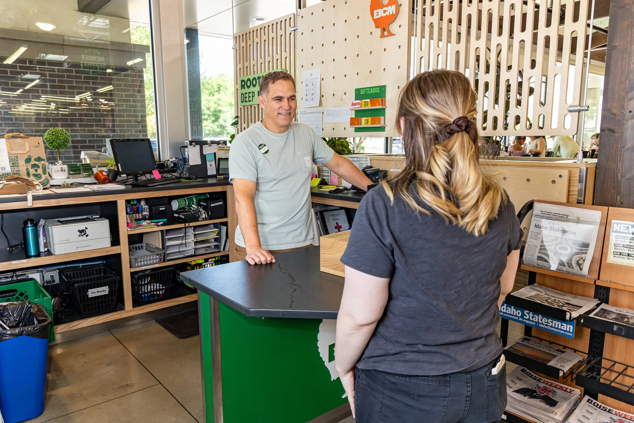 A cashier and a female customer engaging in conversation at the checkout counter inside a retail store. The cashier is smiling and leaning on the counter, while the customer, with her back to the camera, is standing in front of the register. There are various items and magazines on the counter and shelves around.