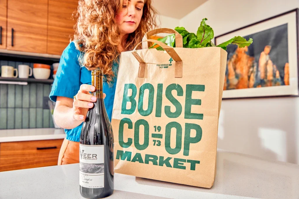 A woman with curly hair and a blue top packing a reusable shopping bag from Boise Co-op Market with green vegetables inside, standing in a kitchen with wooden cabinets and mugs on shelves.