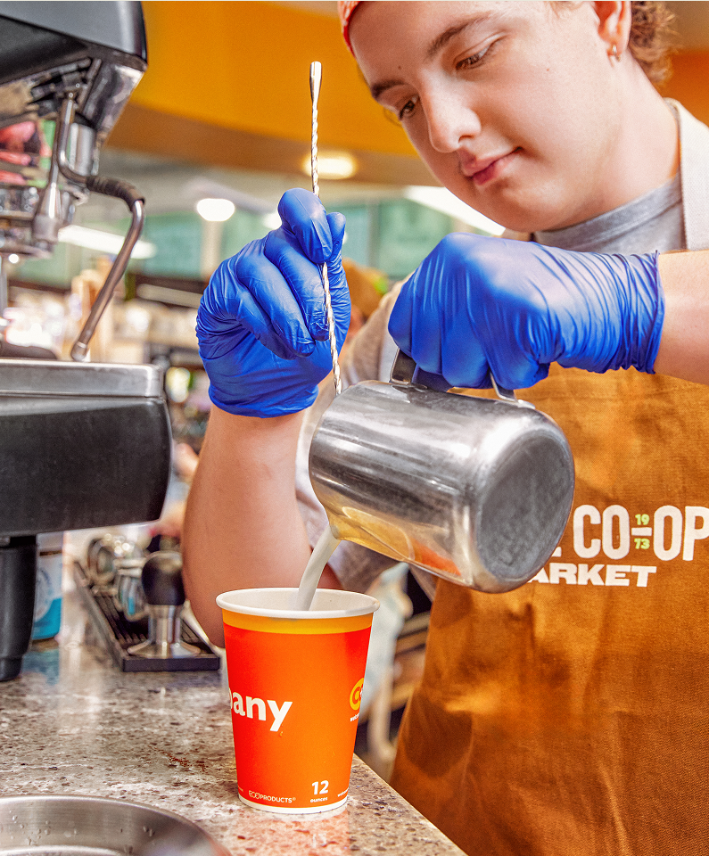 Woman wearing a red hairband, blue gloves, and an orange apron pouring milk into a red and yellow paper cup at a coffee shop, with espresso machine in the background.