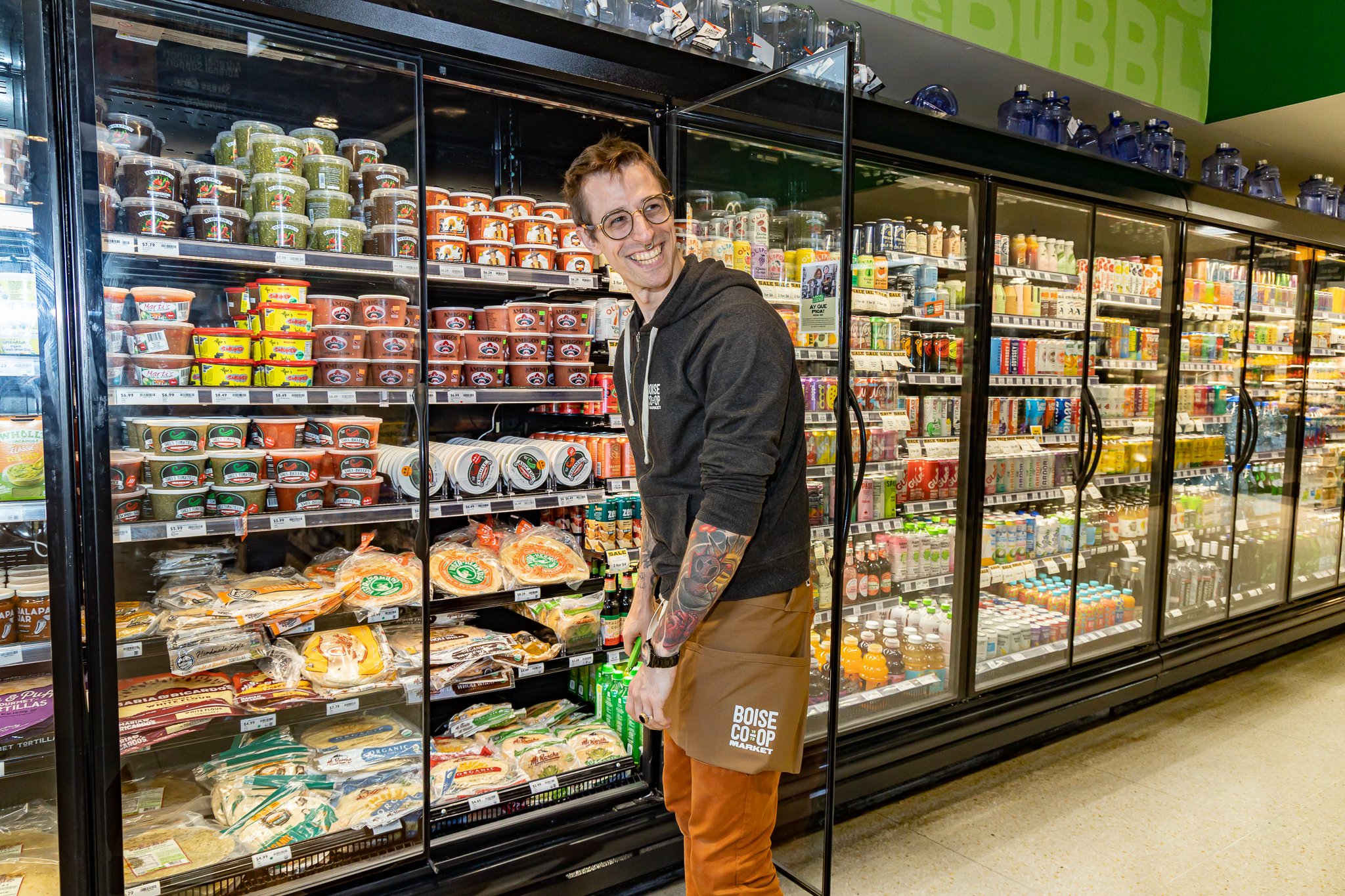 A man with glasses and tattoos on his left arm shopping in a grocery store aisle, smiling while looking towards the right.