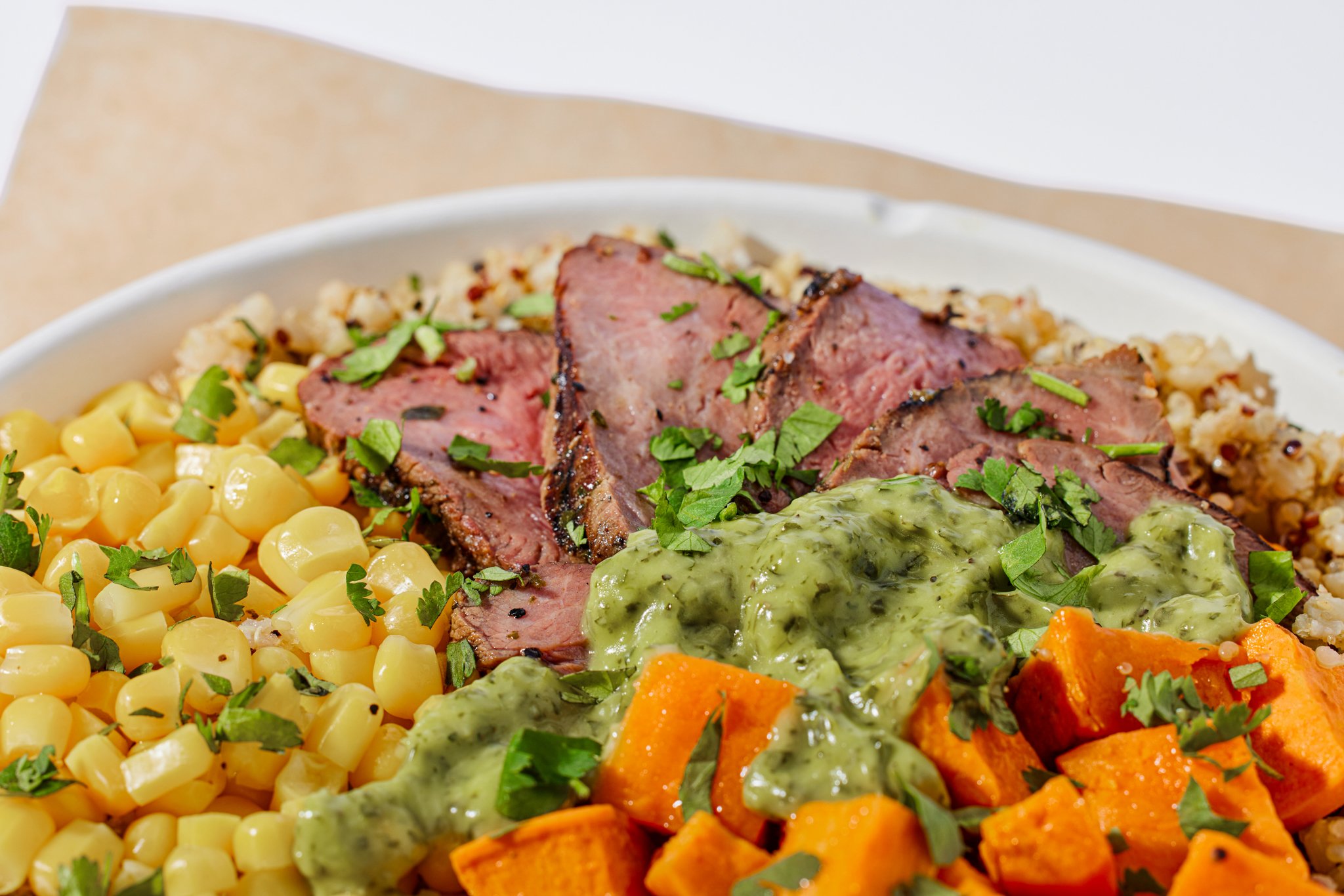 Close-up of a plate with sliced steak topped with cilantro and green sauce, beside corn kernels garnished with herbs, roasted carrots with herbs, and sweet potato cubes.