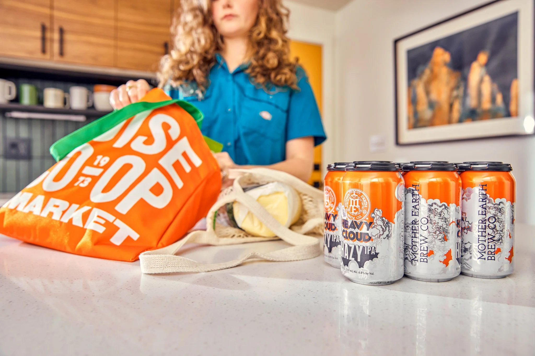 A woman in a blue shirt standing in a kitchen with wooden cabinets in the background. On the counter in front of her are multiple cans of Mother Earth Brewery beer, a tote bag, some sandwiches in a paper bag, and a large orange and white shopping bag.
