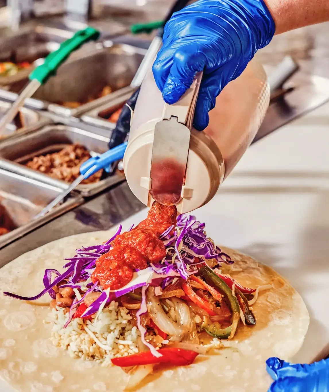 Person with blue gloves adds sauce to a taco filled with shredded vegetables and rice.