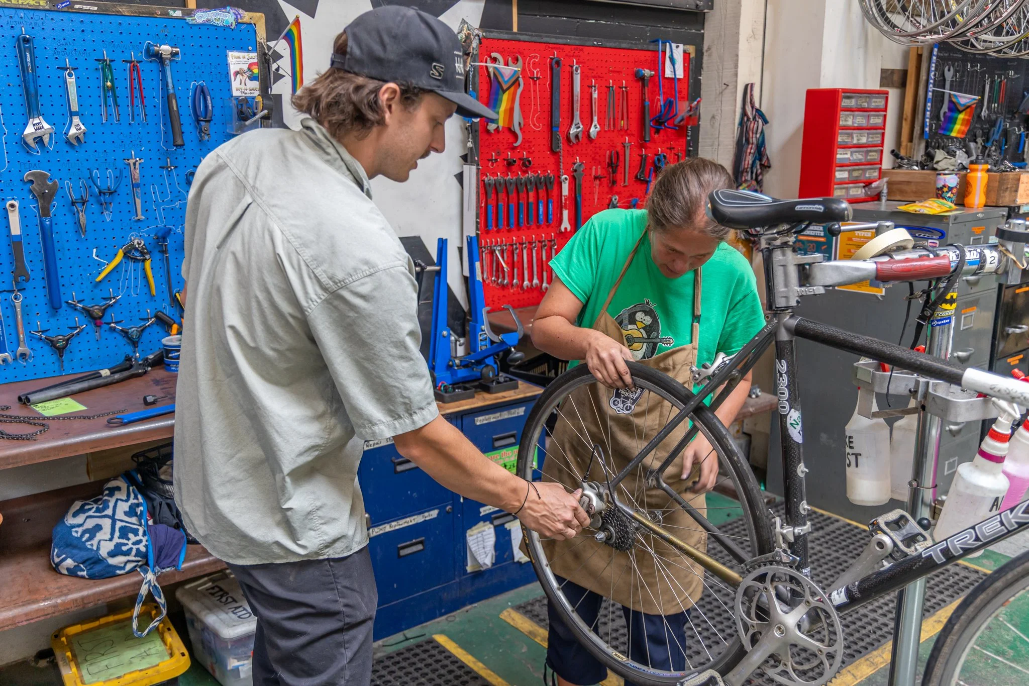 Two people working together on a bicycle in a workshop surrounded by tools and equipment.