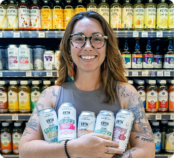 Smiling woman with glasses holding four cans of Earth Organic sparkling water in a grocery store aisle with rows of other canned beverages behind her.