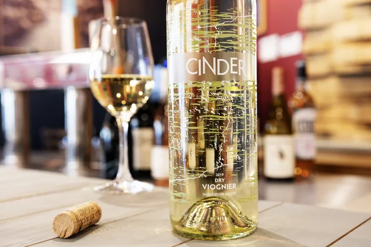 A bottle of Cinder dry viognier wine, with a glass of white wine and a cork on a wooden table, in a setting with shelves and bottles in the background.