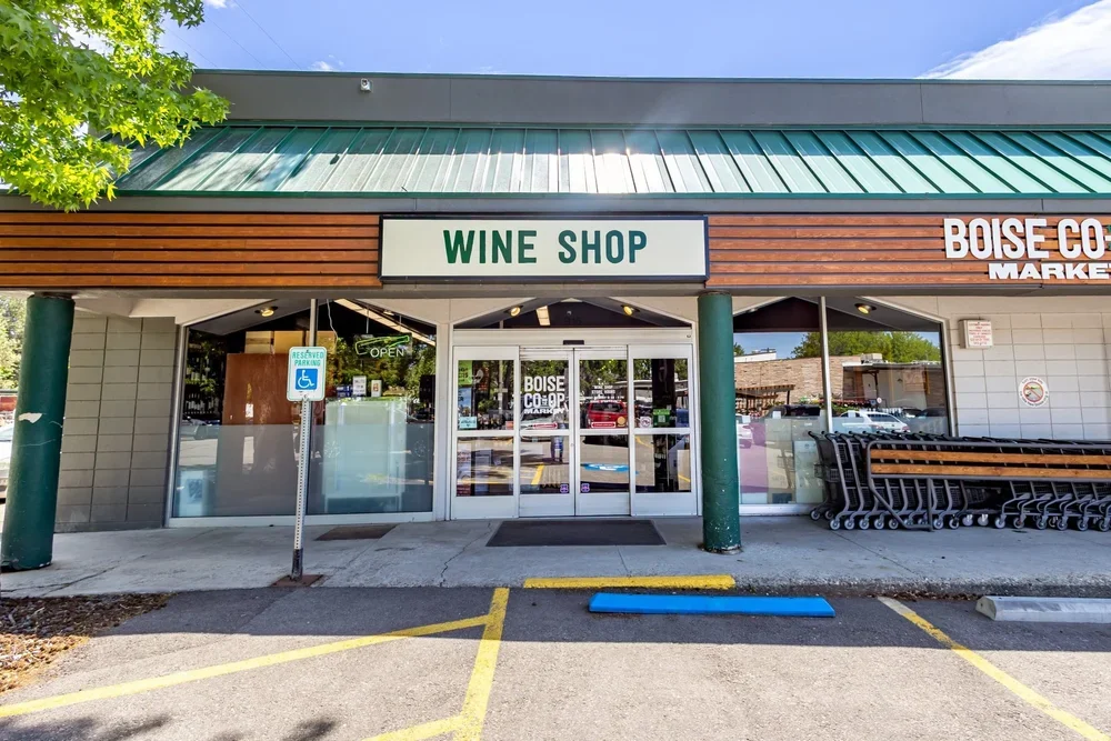 Front of Boise Co-Op market with a sign for a wine shop above the entrance, large glass doors, shopping carts on the right, handicapped parking sign on the left, and a green tree branch and blue sky in the background.