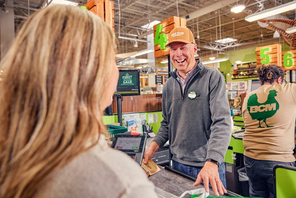 A smiling male cashier at a grocery store checkout counter greets a female customer, with another employee in a BCMS shirt in the background in an aisle with green signage and wooden decoration.
