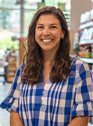 Young woman with long curly hair smiling, wearing a blue and white checkered shirt, inside a bookstore or library.