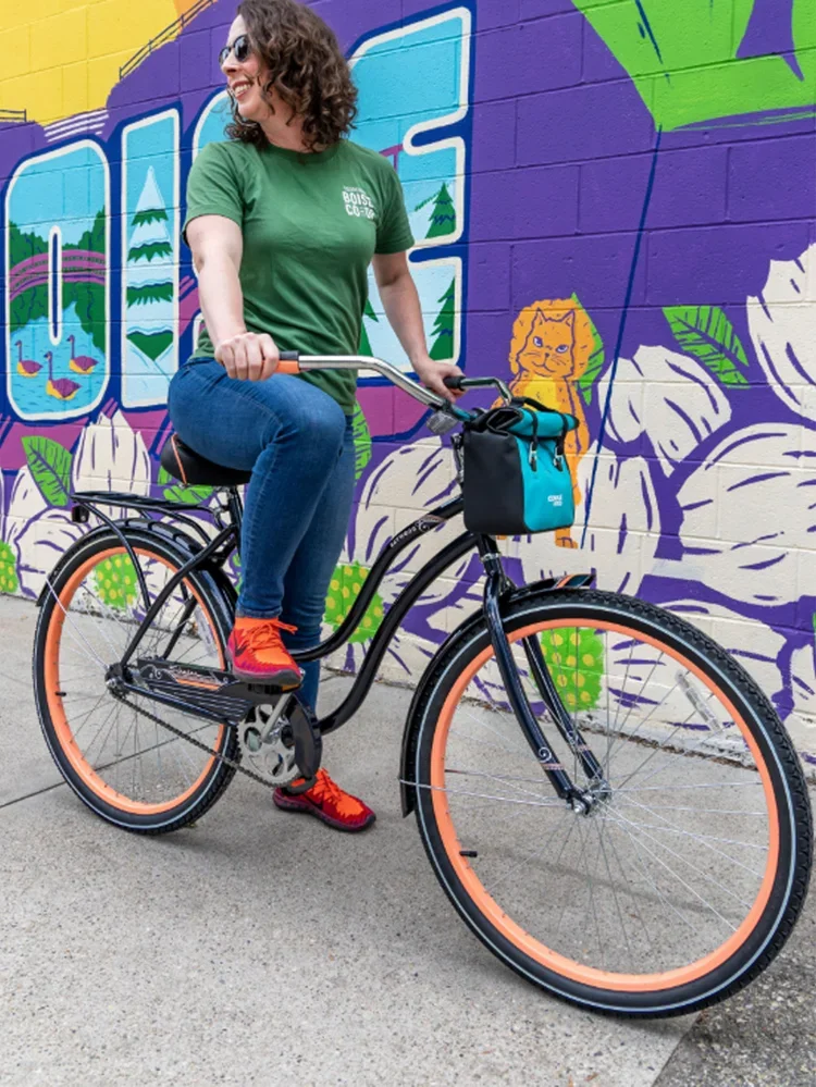 A woman with curly hair and sunglasses, wearing a green T-shirt, blue jeans, and red shoes, is sitting on a black bicycle with peach-colored tires. She is in front of a colorful mural on a purple wall, depicting a lion, plants, and water scenes.