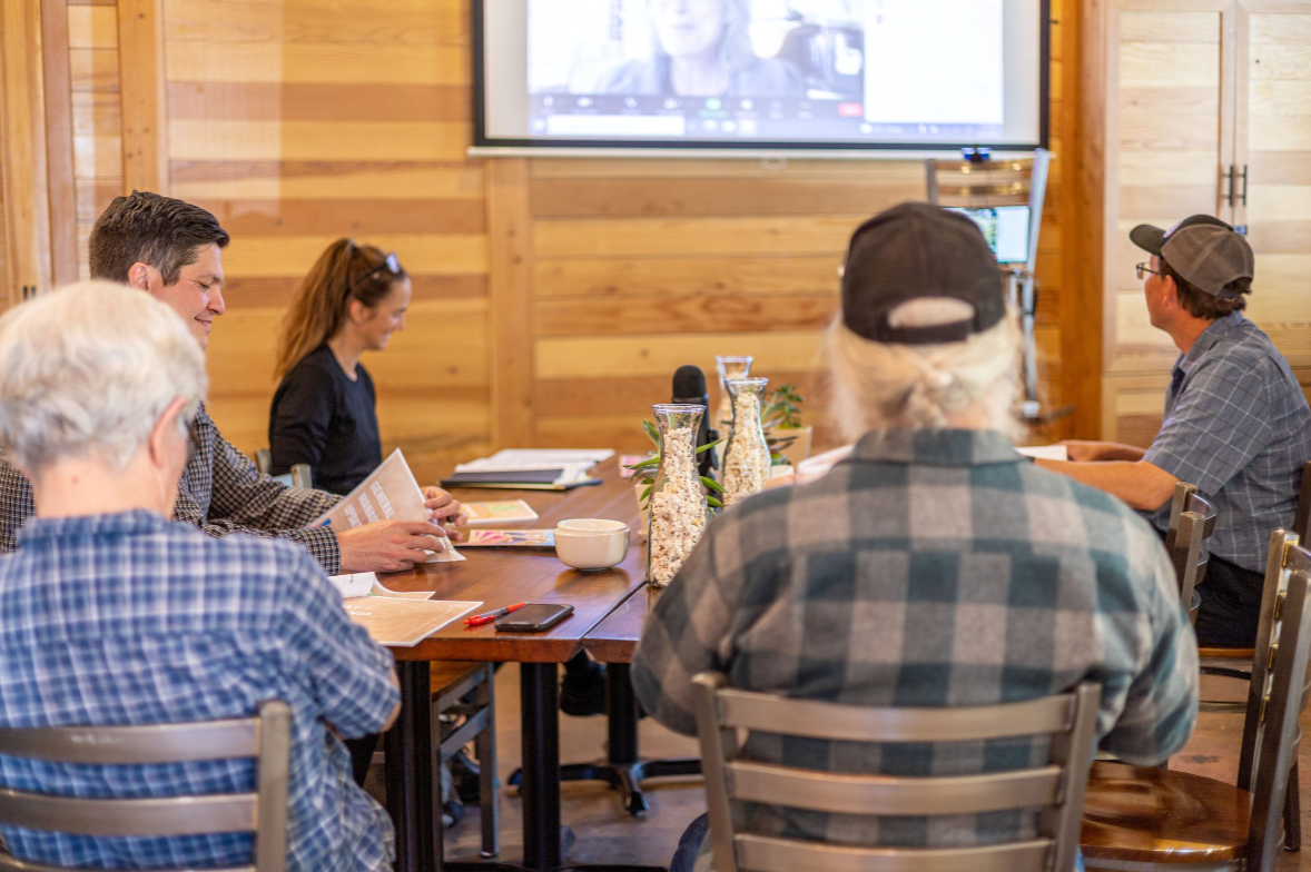 People attending a meeting or workshop in a wooden-paneled room, with a large screen at the front showing a video call and tables with notebooks, drinks, and decorative vases.