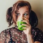 Woman with red hair drinking from a green mug, wearing a black polka dot shirt, headphones on, in a casual setting.