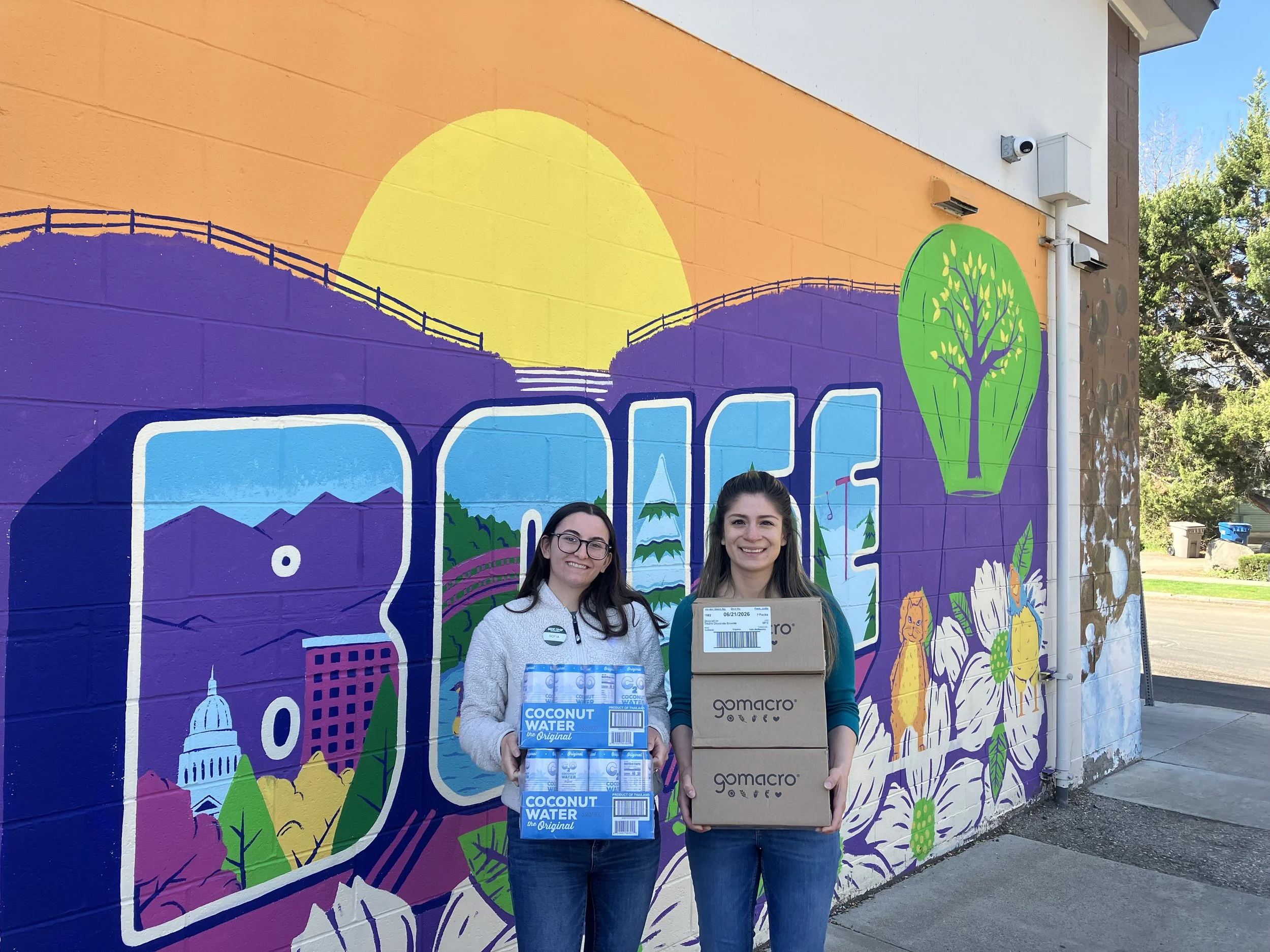 Two women standing in front of a colorful mural holding boxes of coconut water and a cardboard box labeled 'gomacro'.