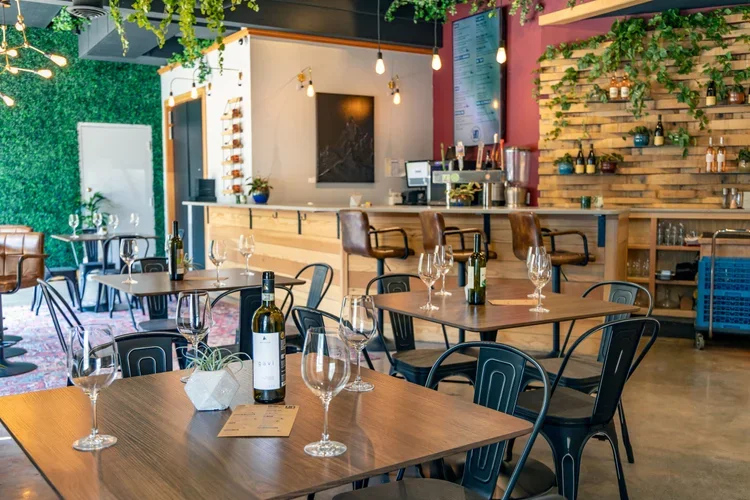 Interior of a modern restaurant or cafe with wooden tables, wine bottles, wine glasses, and a bar with a menu board, surrounded by green plants and colorful decor.