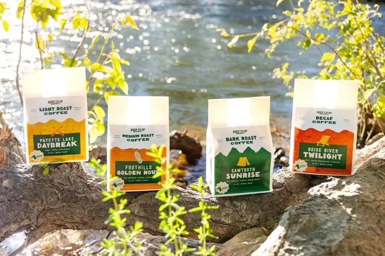 Four bags of Boise Co-op coffee with different labels, set on rocks near a body of water surrounded by greenery.