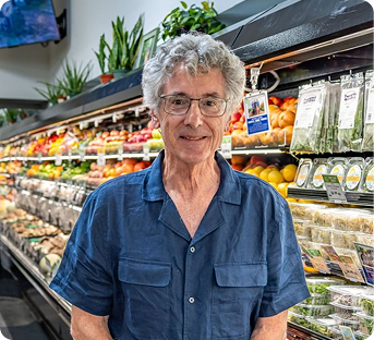 An elderly man with gray curly hair and glasses shopping in a grocery store, standing in front of a produce section with various fruits and vegetables.