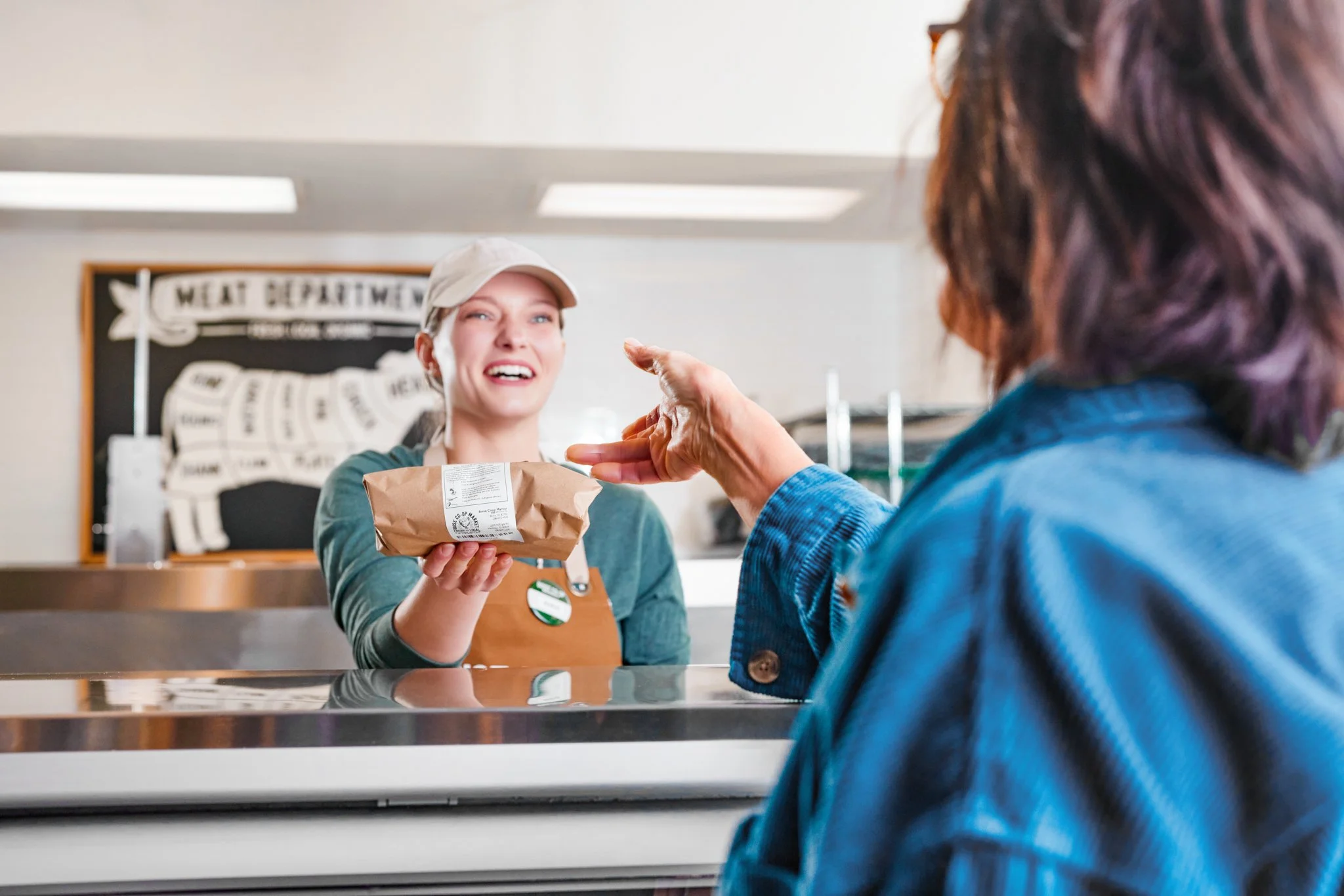 A smiling woman wearing a beige cap and apron hands a brown paper bag to another person in a blue jacket inside a coffee shop.