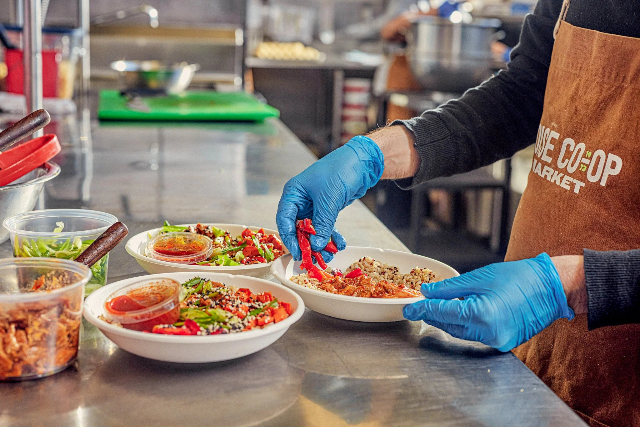 A person wearing a brown apron and blue gloves is preparing food in a kitchen. They are handling a red bell pepper over several bowls filled with different dishes and ingredients, with a green cutting board and kitchen shelves in the background.