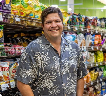 A smiling man wearing a floral shirt standing in a grocery store aisle with snack chips on shelves behind him.