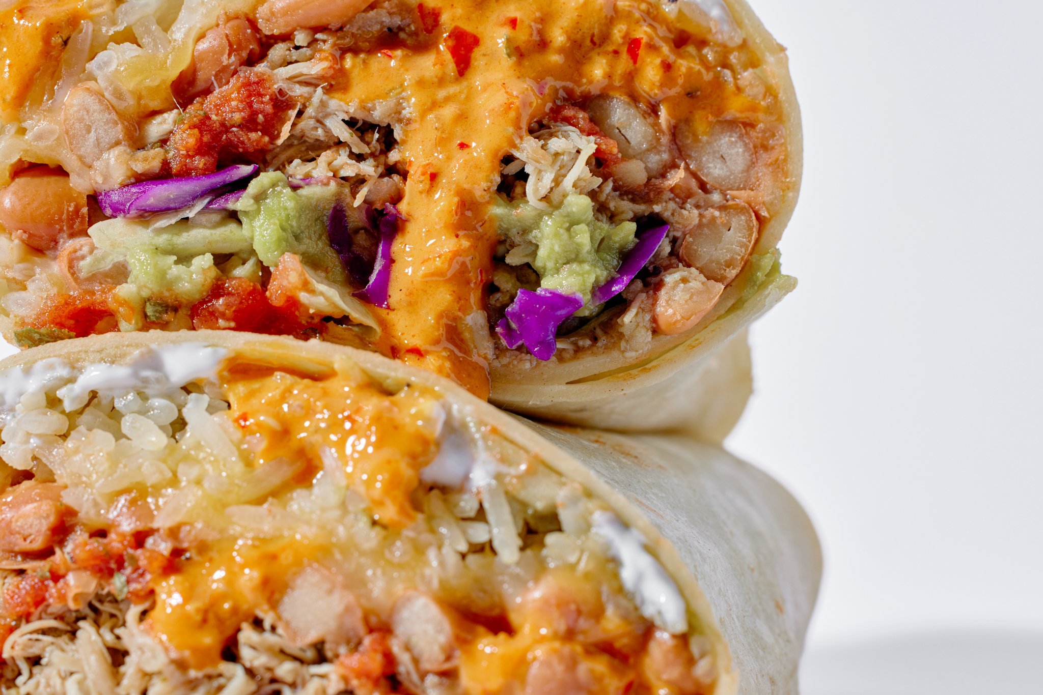 Close-up of a burrito cut in half, showing rice, shredded chicken, lettuce, purple cabbage, tomato, shredded cheese, and orange sauce inside a flour tortilla.