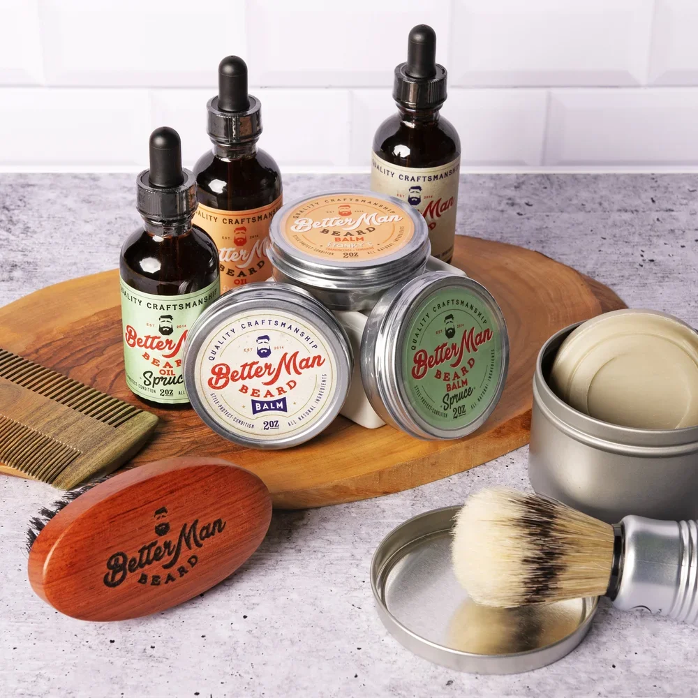 Assorted beard grooming products including beard balm, beard oil, and beard spray bottles on a wooden board with a wooden beard grooming brush and a container of beard balm. A rock with the brand name 'Better Man Beard' is also present.