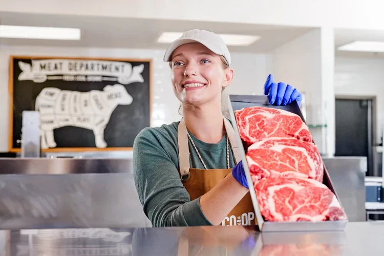 A smiling woman in a white cap and apron holds a tray with three large cuts of raw beef steaks in a butcher shop or meat department.