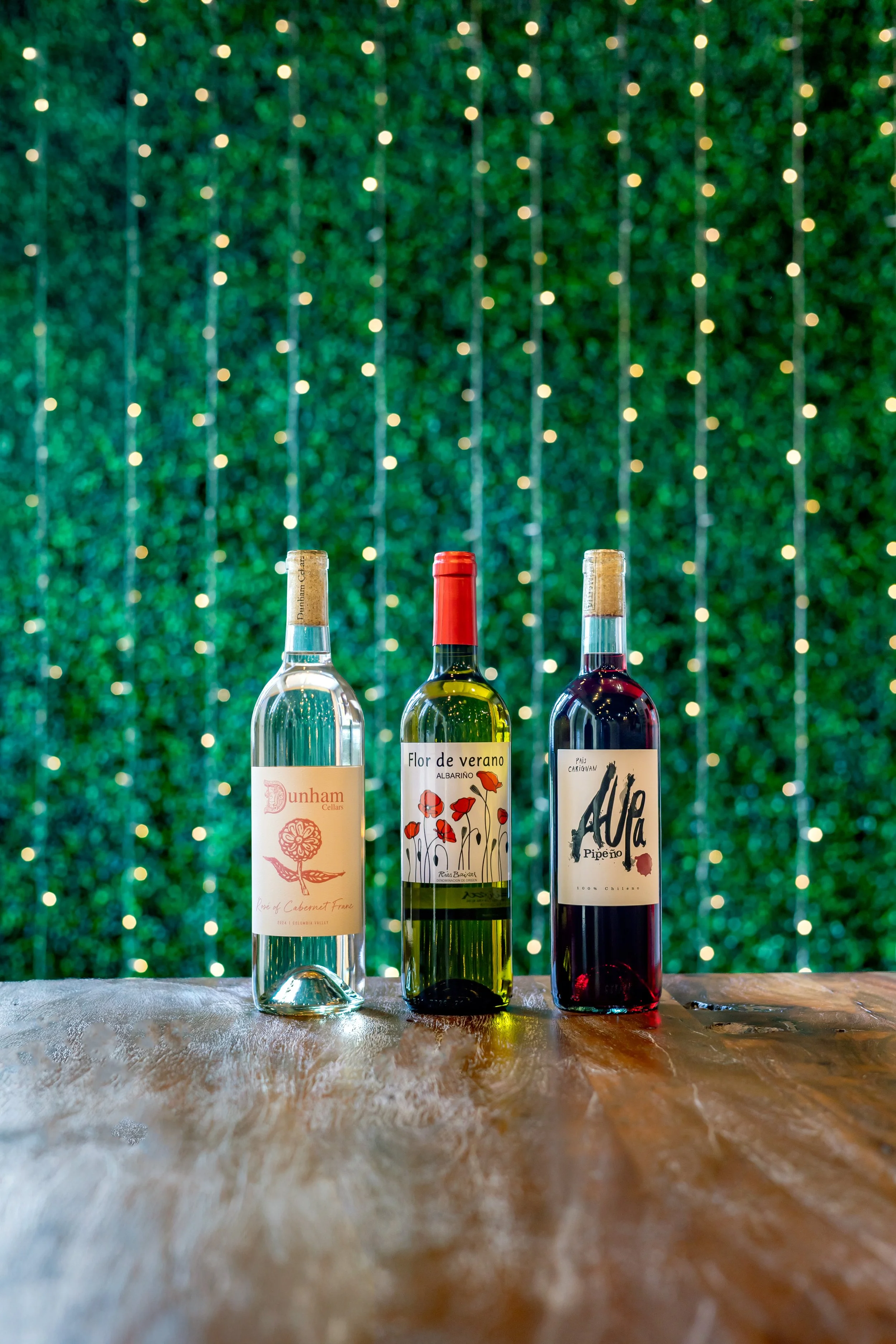 Three bottles of wine on a wooden surface with a blurred green leafy background with string lights.
